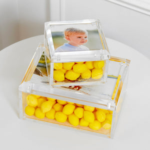 Two Tara Wilson Designs Small Clear Photo Boxes, filled with yellow candies, are stacked on a white surface; the top box displays a child’s photo on the lid—perfect for sharing memories.