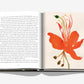 The book AlUla Flora by Assouline is open, showing text on the left page and a colorful illustration of vibrant flora on the right page. The background is white, and the book rests on a white surface.