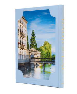 A hardcover book titled Baur Au Lac: A Legacy by the Lake from Assouline, featuring a cover image of a riverside building, trees, and a bridge beneath a blue sky. Inspired by the timeless elegance of Zurich’s iconic luxury hotels, its spine is adorned with “BAUR AU LAC” in gold lettering.