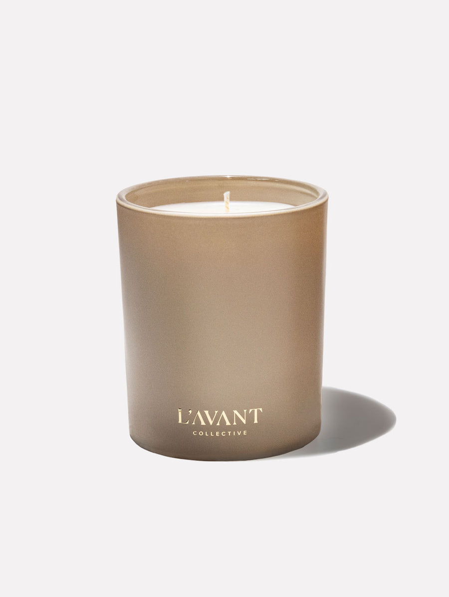 A matte, metallic gold Ambre Santal Collection candle in a cylindrical jar with the L’Avant Collective logo printed in white, evoking the essence of a luxury perfume.