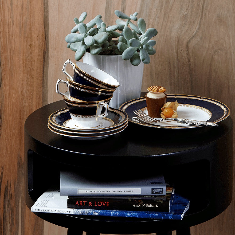 Three stacked Ashbourne Navy teacups with matching saucers and plates from Royal Crown Derby rest on a round black table beside a potted succulent, with pastries and books nearby, showcasing refined British craftsmanship.