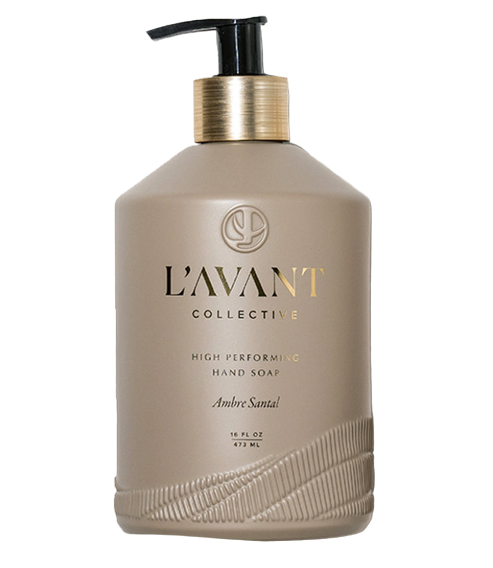 A 16 fl oz (473 ml) beige bottle of L'Avant Collective Ambre Santal Collection Hand Soap features a black pump, gold label, and a luxury blend of cardamom, amber, and cedar.