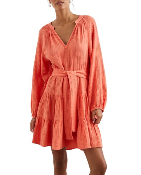 A woman in a long-sleeve, coral pleated Rails Aureta dress with a v-neckline and tied waist, standing against a white background.