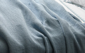 The Matouk Chatham Blanket in Hazy Blue, featuring a textured cotton waffle design and made in Portugal, is draped over a bed with a white sheet edged in a blue wavy pattern.