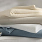 Four folded Chatham Blankets by Matouk in ivory, beige, white, and blue are stacked on a light surface beside a bowl with white flowers. Each blanket is 100% cotton, crafted in Portugal.