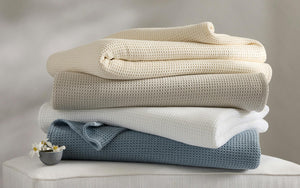 A stack of four Matouk Chatham Blankets in cream, beige, white, and blue—crafted in Portugal from 100% cotton waffle weave—rests on a white surface beside a small bowl of flowers.