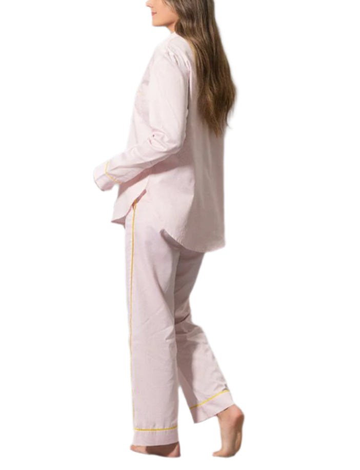 A person with long hair is wearing the Matouk Jasper Pajama Set in light pink cotton sateen with yellow piping, standing sideways and barefoot against a white background.