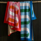 Two vibrant Matouk Ribbon Beach Towels with bold, curved patterns in red/pink and blue/green hang over a wooden branch against a dark ribbed wall. Made from soft cotton terry, they add stylish flair to any setting.