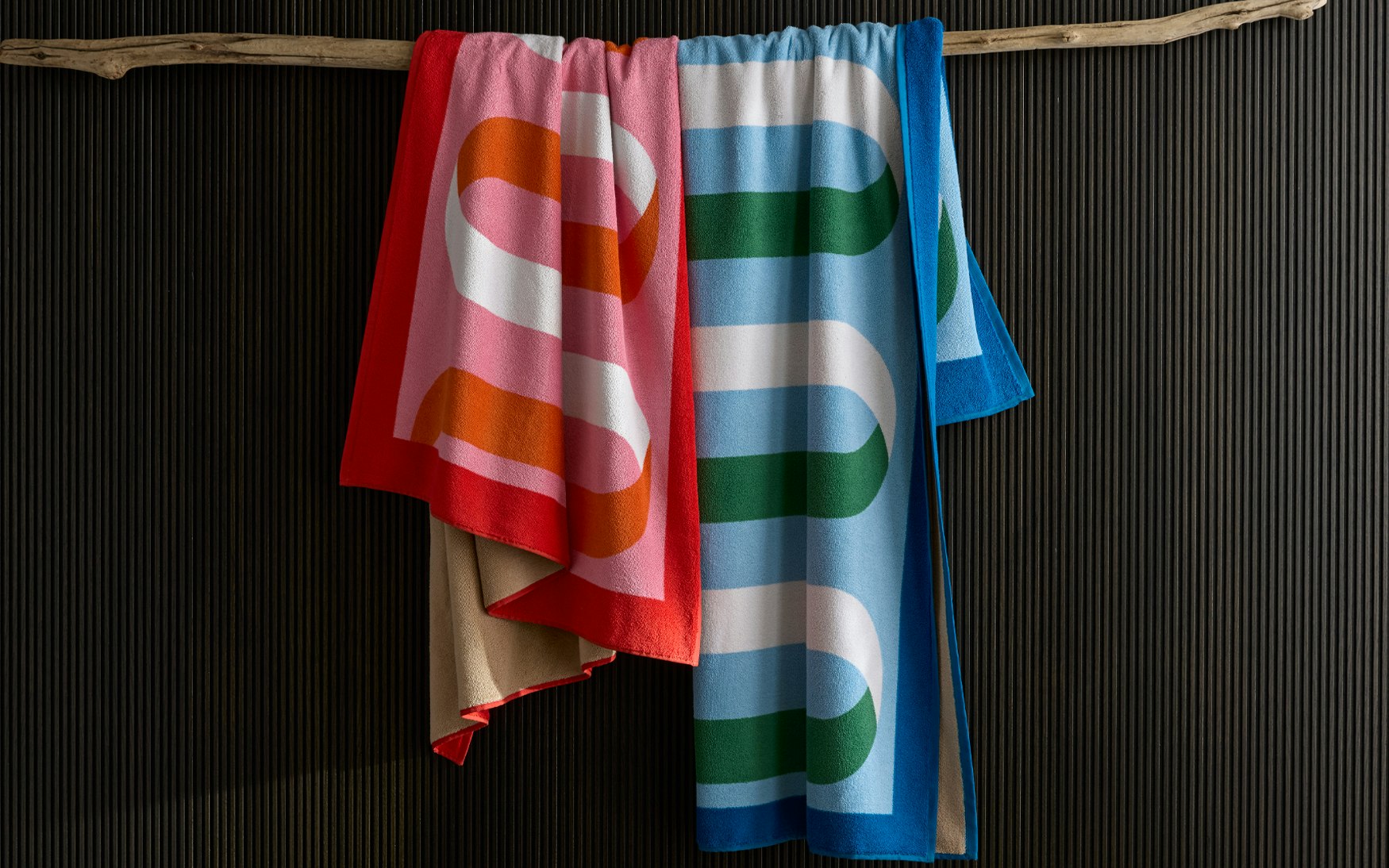 Two vibrant Matouk Ribbon Beach Towels with bold, curved patterns in red/pink and blue/green hang over a wooden branch against a dark ribbed wall. Made from soft cotton terry, they add stylish flair to any setting.