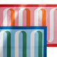Two Matouk Ribbon Beach Towels—one in red, pink, orange, and white; the other in blue, green, teal, and white. The blue towel overlaps the red one and both feature arched stripe patterns on vibrant cotton terry.
