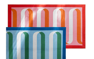 Two Matouk Ribbon Beach Towels—one in red, pink, orange, and white; the other in blue, green, teal, and white. The blue towel overlaps the red one and both feature arched stripe patterns on vibrant cotton terry.