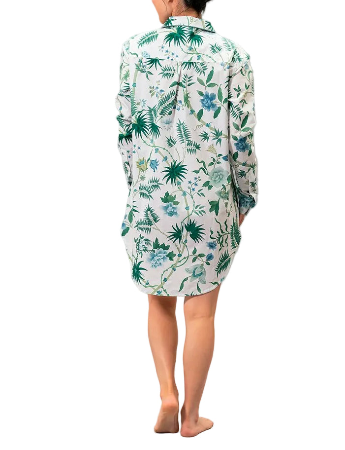 A person stands barefoot, facing away, wearing the Matouk Luca Palmyra Emerald Sleep Shirt—an oversized, long white pajama top featuring a green and blue floral leafy pattern.