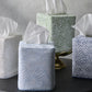 Four Matouk Duma Diamond Dune Tissue Box Covers in white, light blue, green, and dark blue—each with a subtle geometric pattern—inspire an elegant look as tissues protrude from each on a dark surface.