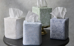 Four Matouk Duma Diamond Dune Tissue Box Covers in white, light blue, green, and dark blue—each with a subtle geometric pattern—inspire an elegant look as tissues protrude from each on a dark surface.