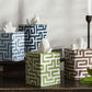 Four Matouk Gabriel Tissue Box Cover Collection boxes with blue, green, and brown geometric motifs sit near two tall white candles and a green leaf, bringing stylish flair to your bathroom decor.