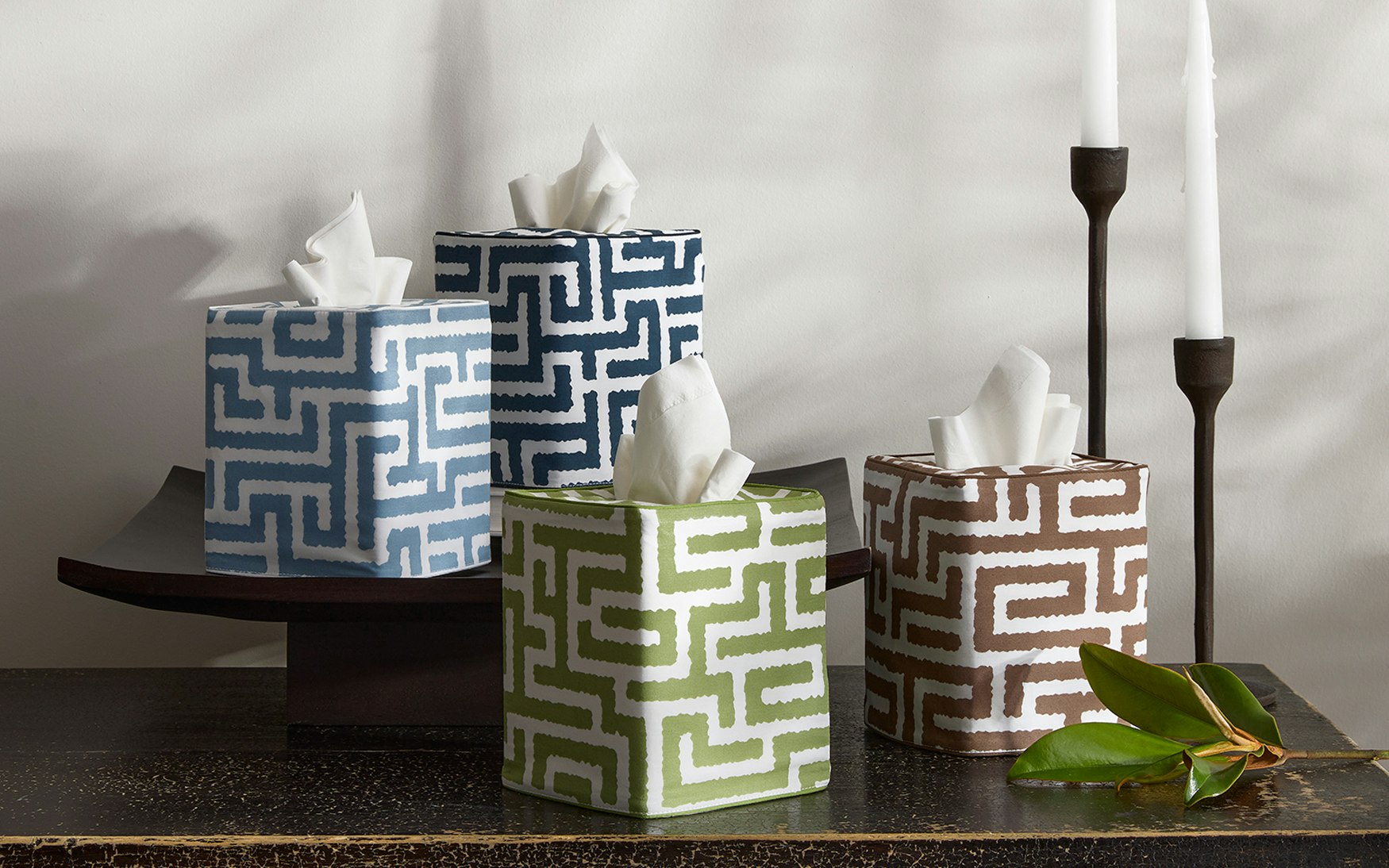 Four Matouk Gabriel Tissue Box Cover Collection boxes with blue, green, and brown geometric motifs sit near two tall white candles and a green leaf, bringing stylish flair to your bathroom decor.