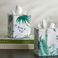 Two Matouk Palmyra Tissue Box Covers in Emerald, featuring botanical prints, bring island-inspired decor to a dark table; one sits atop a stack of three books.