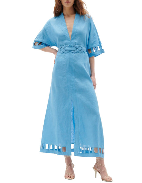 A woman is wearing the Figue Adalaide Dress, a long blue kaftan-style dress by Figue, featuring short, wide sleeves and a Love Knot belt. The 100% linen dress incorporates elegant cutout designs near the bottom and on the sleeves. She has paired it with beige heels.