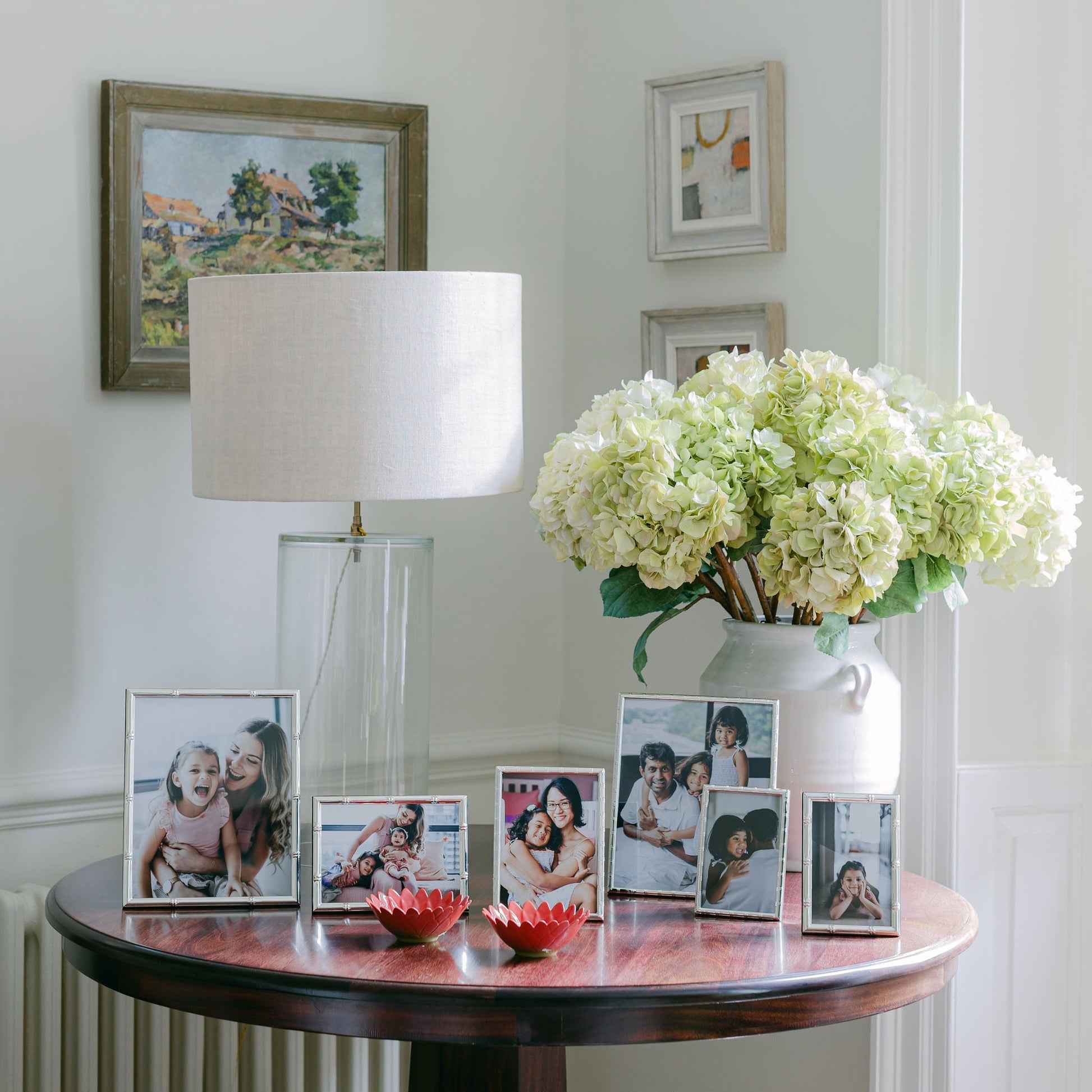 A round wood table with Addison Ross’s Cane Silver Frame Collection displays family photos, a lamp, two red bowls, and a white hydrangea vase, set against a white wall decorated with framed artwork.