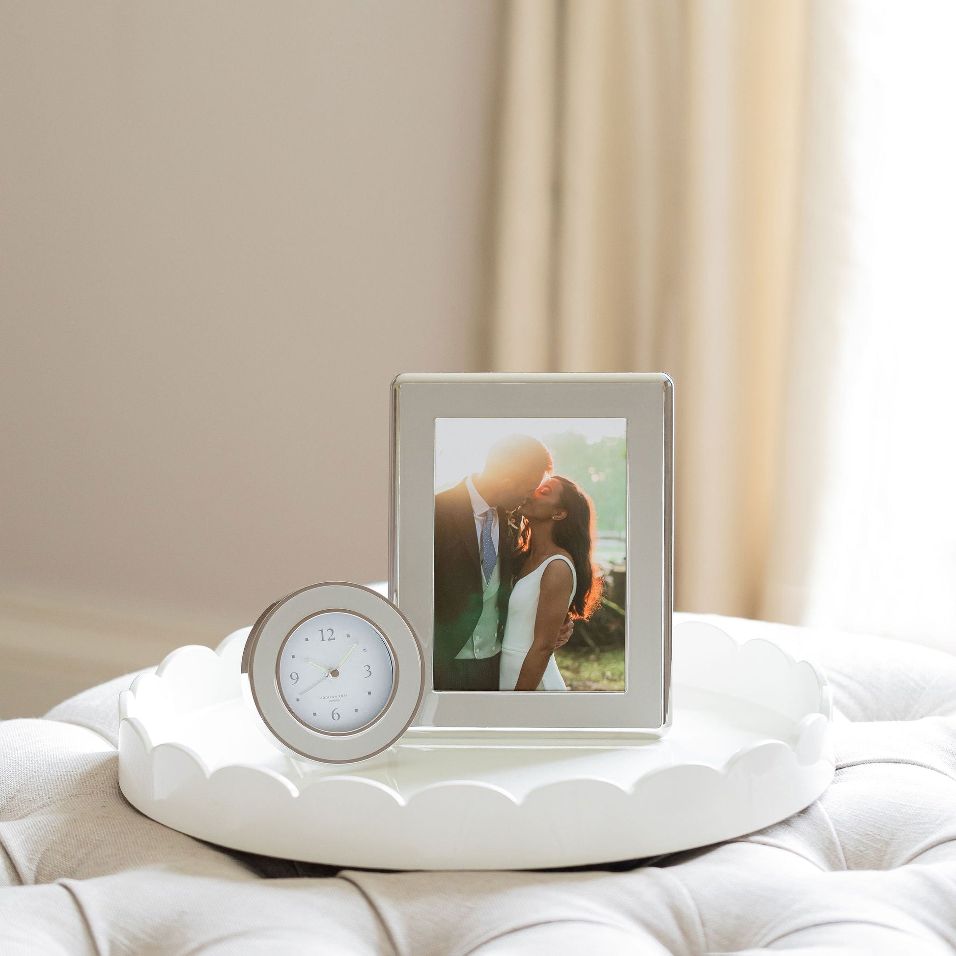 A framed photo and the Addison Ross Round Silver Alarm Clock rest on a white tray atop a cushioned surface, with soft curtains in the background—an inviting, cozy setup for your bedside table.