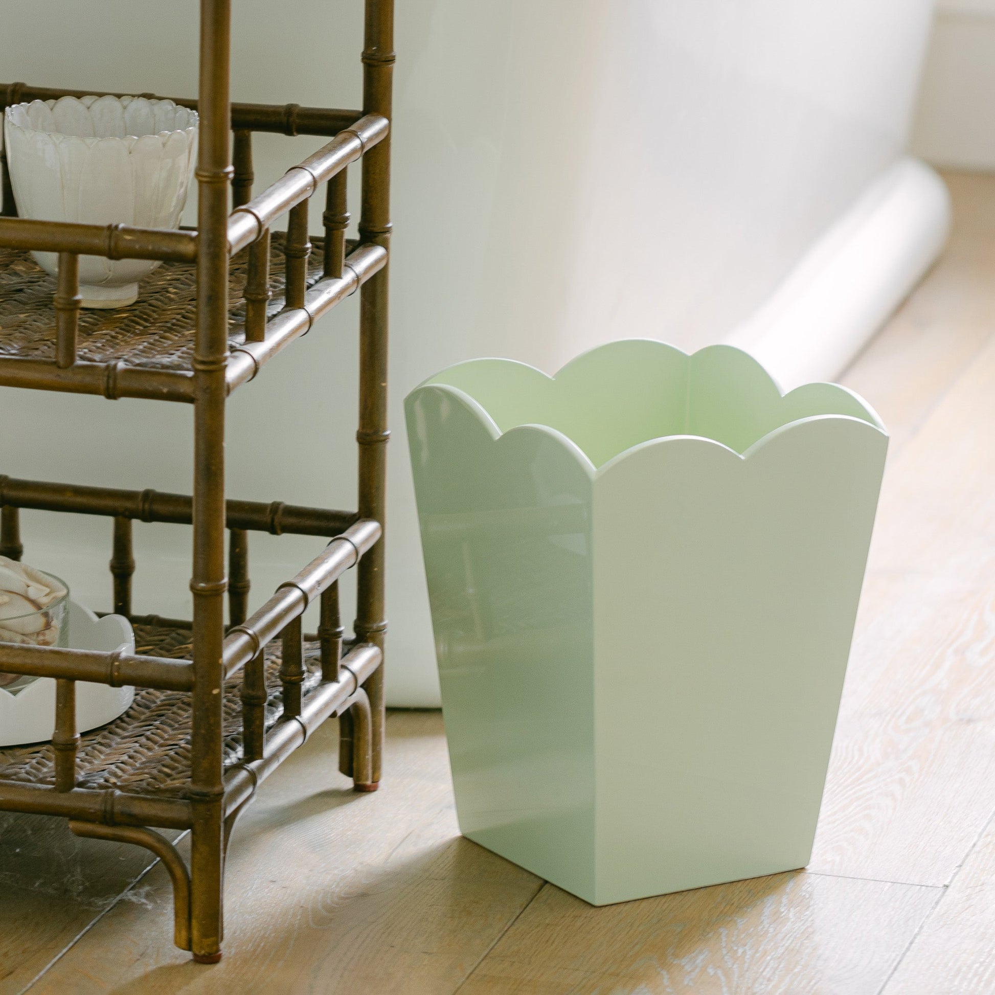 The pale green scalloped bin from the Addison Ross Scalloped Lacquer Waste Bin Collection by Addison Ross sits elegantly next to a basket weave wooden shelf on a wooden floor.
