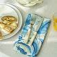 A plate with sliced bread, a fork and knife from the Addison Ross Wave Flatware Collection on a blue-patterned napkin, a glass of orange juice, and a spoon with saucer rest on a white tablecloth.