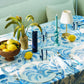A table set with the Addison Ross Palm Beach Tablecloth in blue and white, accompanied by plates, glasses, a vase of grass, fresh lemons, a basket of lemons, candles, and a small lamp.