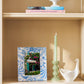 A bookshelf displays an Addison Ross Palm Beach Frame Collection photo frame with a blue pattern, a green candle in a holder, a white decorative bird, and two stacked books on the top shelf.