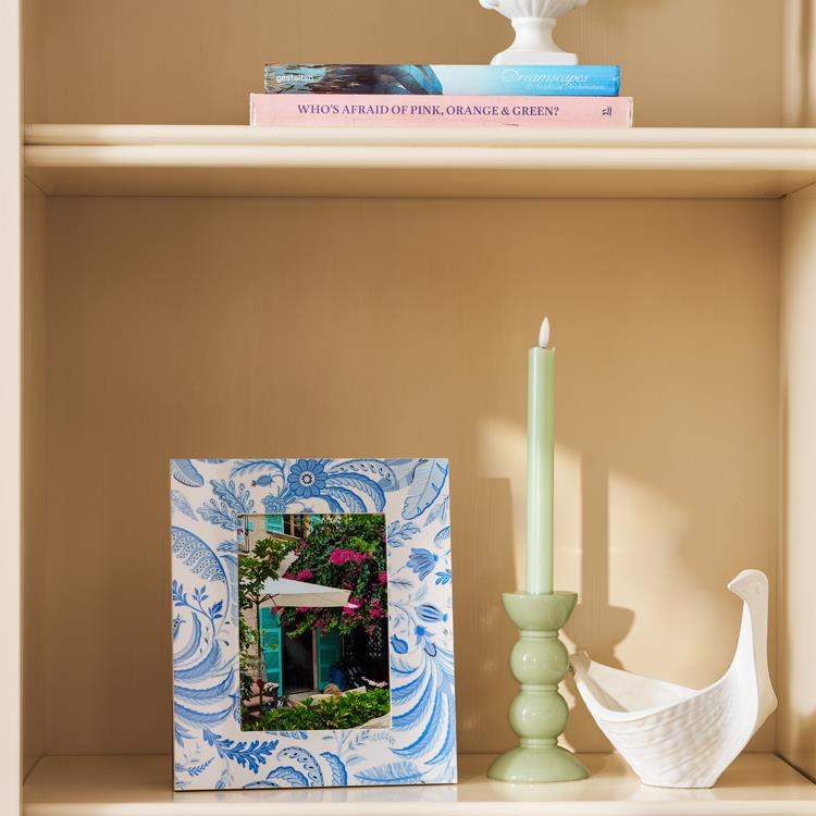 A bookshelf displays an Addison Ross Palm Beach Frame Collection photo frame with a blue pattern, a green candle in a holder, a white decorative bird, and two stacked books on the top shelf.