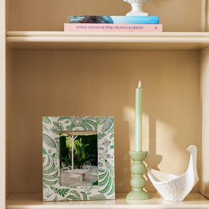 A wooden shelf displays a pink book, an Addison Ross Palm Beach Frame Collection photo frame with an outdoor photo, a green candle in a holder, and a white ceramic bird-shaped dish.