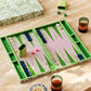 The Addison Ross Palm Beach Backgammon Collection features an open luxury set with green, white, and navy pieces, two pink dice, a green dice cup, leather-and-suede shakers, and a patterned box on a beige surface.