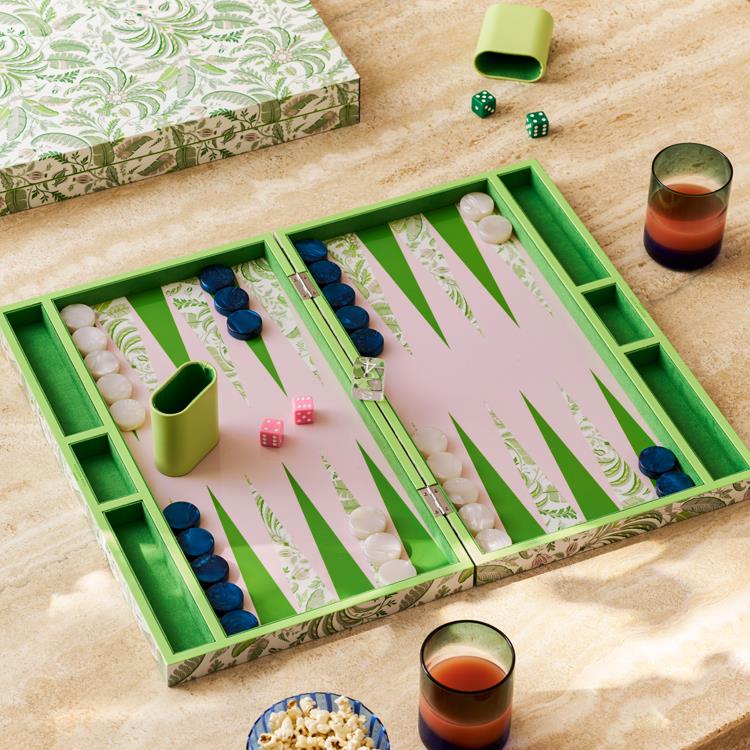 The Addison Ross Palm Beach Backgammon Collection features an open luxury set with green, white, and navy pieces, two pink dice, a green dice cup, leather-and-suede shakers, and a patterned box on a beige surface.