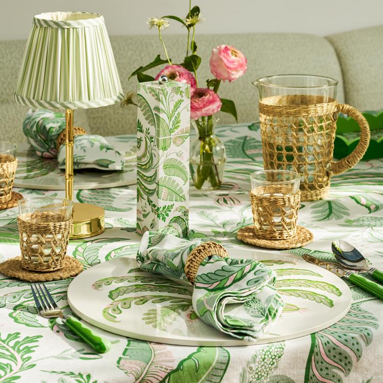 A table set with green and white botanical-themed tableware, wicker glass holders, a glass pitcher, a small lamp, a pink floral vase, and Addison Ross Palm Beach Salt and Pepper Mill Collection made from sustainably sourced rubberwood.