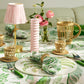A table set with an Addison Ross Palm Beach Tablecloth, featuring leaf patterns, wicker-wrapped glasses and pitcher, pink flowers, a pink candle, and a small lamp with a pink shade.