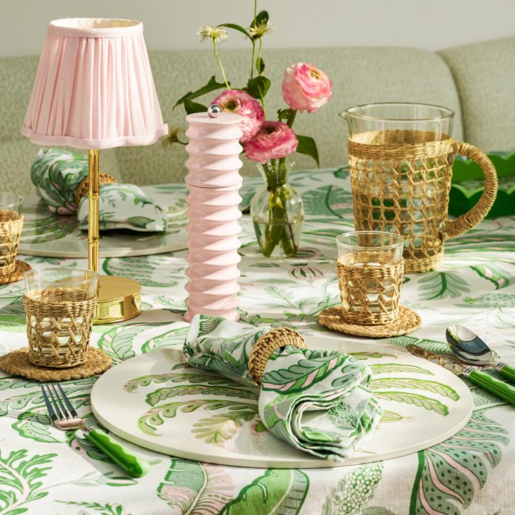 A table set with an Addison Ross Palm Beach Tablecloth, featuring leaf patterns, wicker-wrapped glasses and pitcher, pink flowers, a pink candle, and a small lamp with a pink shade.
