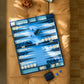 The Addison Ross Palm Beach Backgammon Collection, featuring a lacquered exterior, white and dark blue pieces, dice, a dice cup, mother-of-pearl chips, sits on a tan cushion with a tray of pretzels nearby.