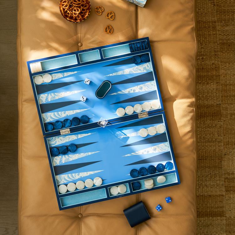 The Addison Ross Palm Beach Backgammon Collection, featuring a lacquered exterior, white and dark blue pieces, dice, a dice cup, mother-of-pearl chips, sits on a tan cushion with a tray of pretzels nearby.