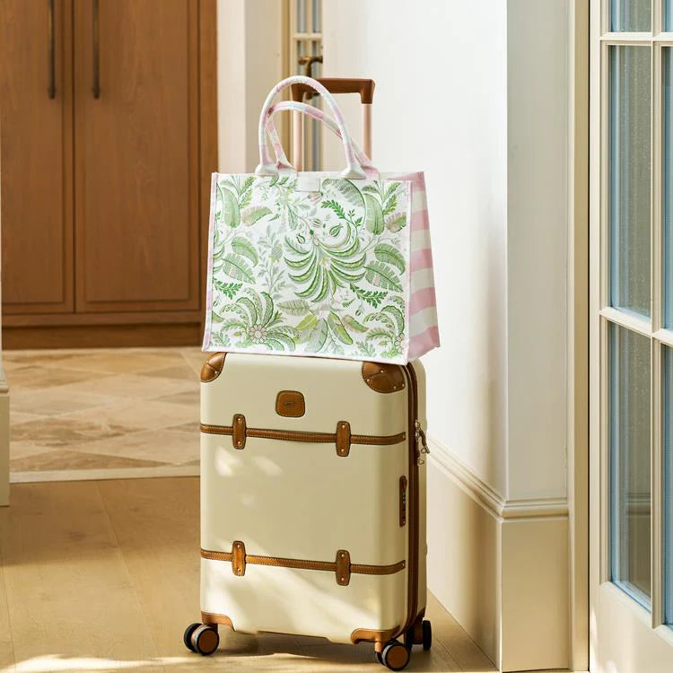 A cream-colored suitcase with brown trim and wheels stands in a hallway, topped with the Addison Ross Palm Beach Tote Bag featuring a chic green and white pattern.