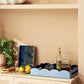 A Scalloped Tray in Pale Denim & Navy by Addison Ross, styled with glassware, a decanter, and a bottle, sits atop a beige cabinet. Lemons, limes, a plant, and a serving board are also arranged against the beige wall.