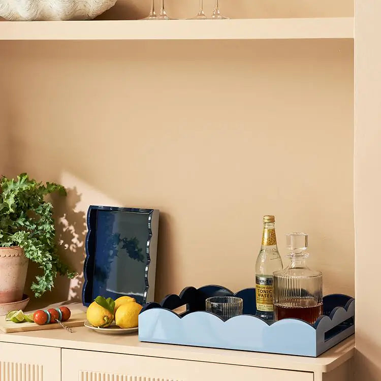 A Scalloped Tray in Pale Denim & Navy by Addison Ross, styled with glassware, a decanter, and a bottle, sits atop a beige cabinet. Lemons, limes, a plant, and a serving board are also arranged against the beige wall.
