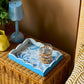 A wicker table styled in Palm Beach decor features a blue lamp, yellow vase, and an Addison Ross Palm Beach Scallop Tray from the collection, holding a glass of water in a wicker holder.