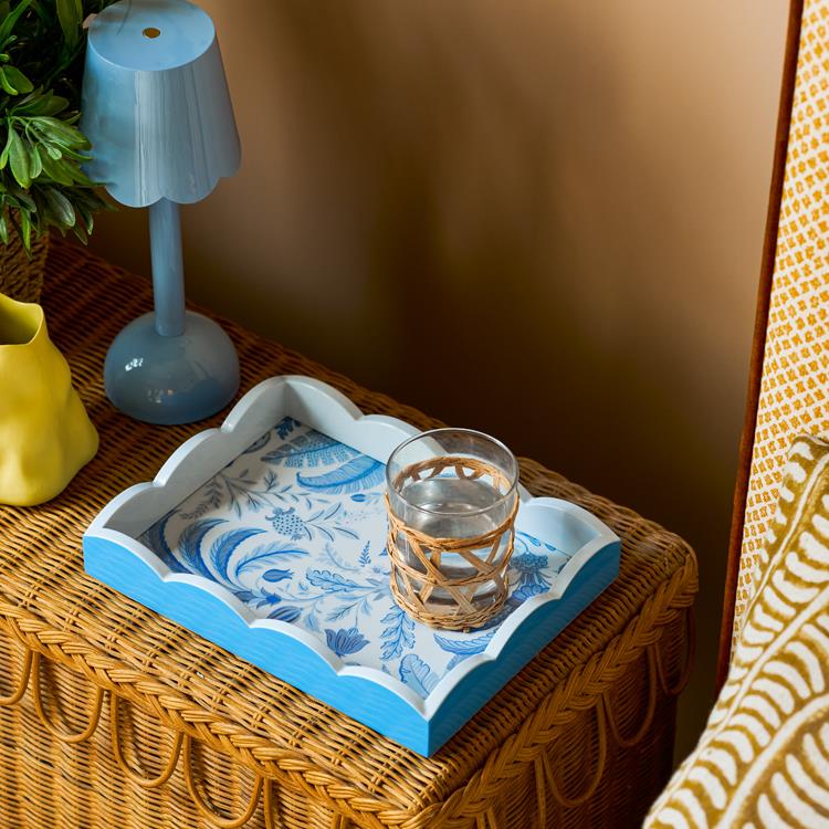 A wicker table styled in Palm Beach decor features a blue lamp, yellow vase, and an Addison Ross Palm Beach Scallop Tray from the collection, holding a glass of water in a wicker holder.