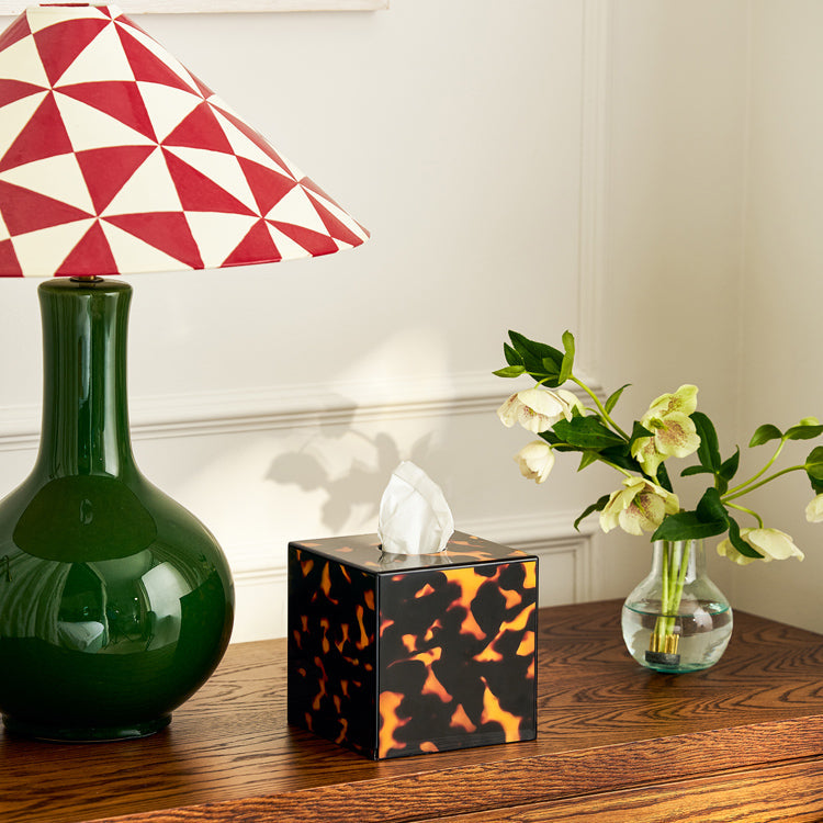 A green lamp with a red and white geometric shade, an Addison Ross Lacquer Tissue Box, and a glass vase with white flowers on a wooden table.