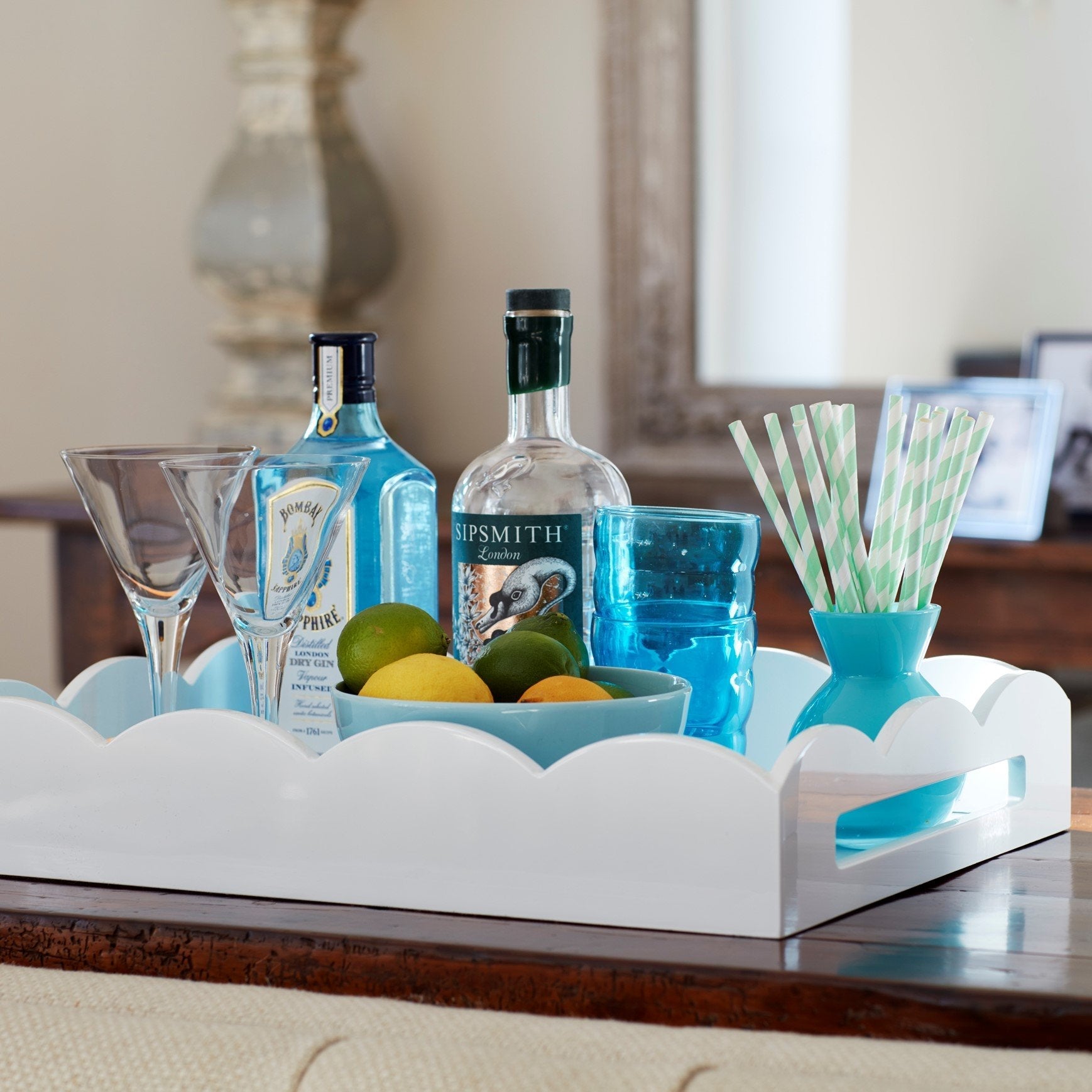 The Scalloped White Lacquer Tray Collection by Addison Ross holds two gin bottles, two cocktail glasses, a bowl of limes and lemons, a blue glass, and a striped straw container.