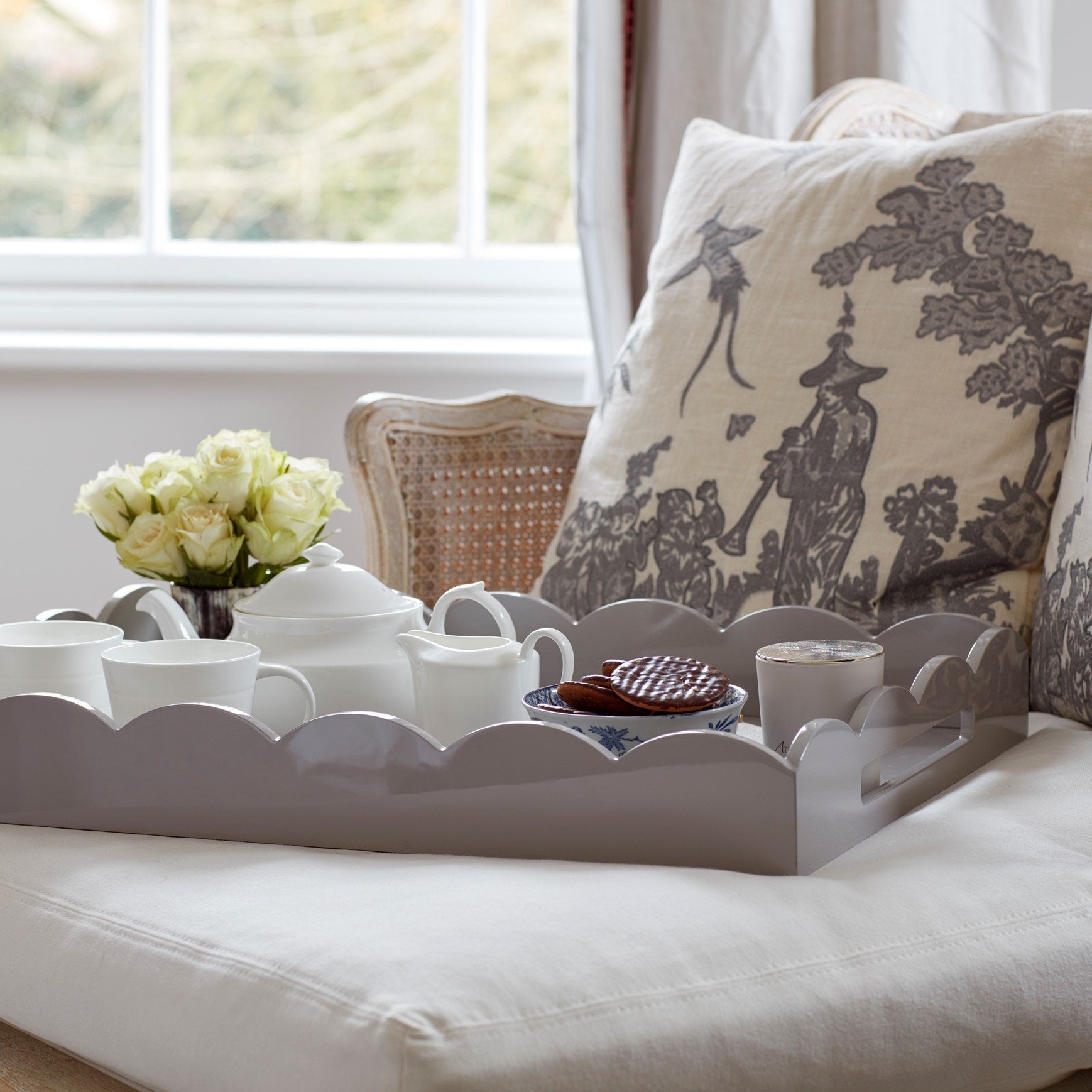 The Addison Ross Scalloped Chiffon Lacquer Tray holds a teapot, cups, saucers, cookies, and a vase of white roses on a cushioned chair with decorative pillows by the window.