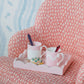 An Addison Ross Scalloped Edge Lacquer Tray in Pale Pink, featuring two mugs, two spoons, and a small decorative turtle, rests on a pink patterned chair.
