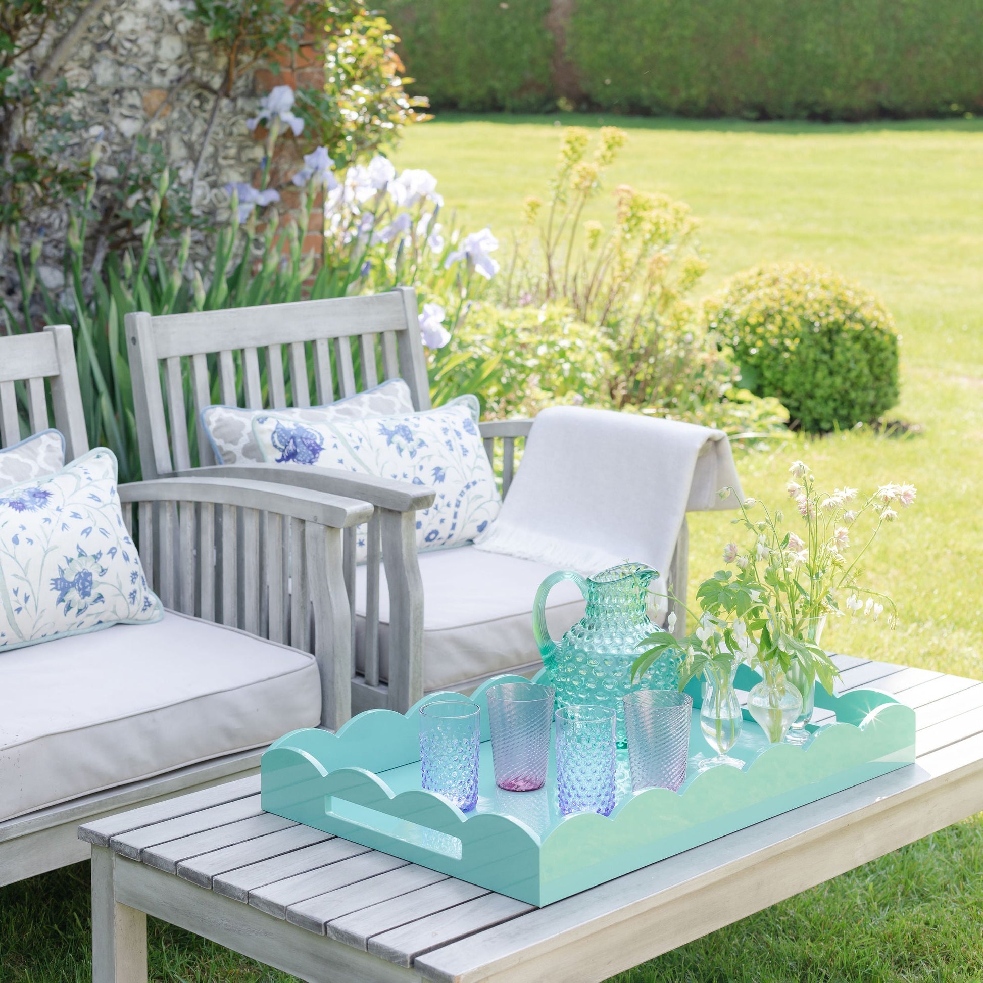 A white wooden bench and table sit in a garden. On the table is an Addison Ross Scalloped Turquoise Lacquer Tray Collection with glassware, a pitcher, and a vase of flowers. Cushions and a throw add comfort to the bench.