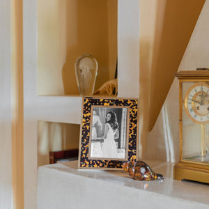 A Windsor Faux Tortoiseshell and Silver Frame by Addison Ross (5 x 7), featuring a black-and-white photo of a woman in a dress, sits on a white shelf beside a glass figurine and a gold clock.