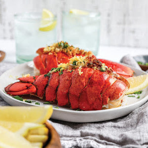 Two lobster tails topped with herbs and breadcrumbs are elegantly arranged on a white plate beside fresh lemon wedges—an ideal healthy seafood meal featured in Coastal Kitchen by Simon & Schuster. In the background, two glasses of water with lemon slices add a refreshing touch to this inviting coastal living setting.