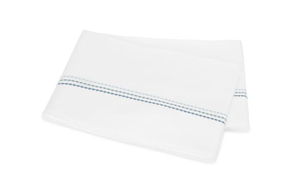 Displayed on a plain white background, the folded white bedsheet from the Matouk Amara Triple Pearl Bedding Collection features an elegant embroidered design of two parallel dotted blue lines near the edge and is crafted from luxurious Amara sateen.